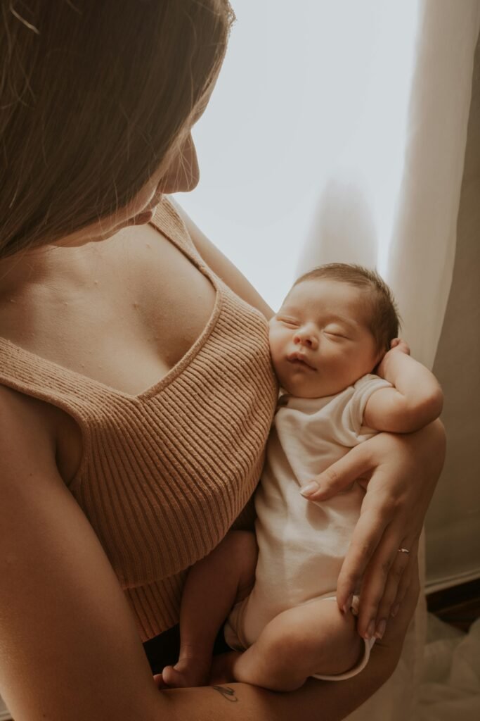 pexels photo 32447392 32447392 Mother lovingly holding her sleeping newborn child by a window with soft lighting.