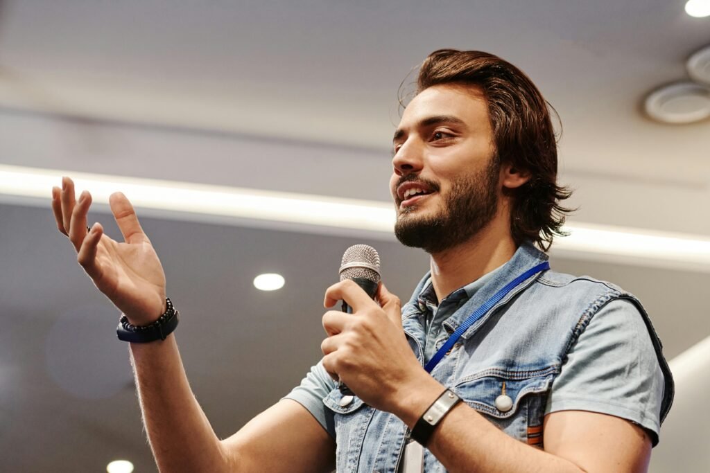 pexels photo 8348462 8348462 Young man with microphone speaking confidently at an indoor conference, gesturing with a smile.